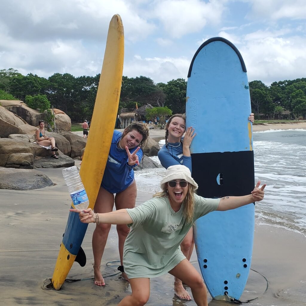 Three girls smiling and enjoying themselves after a surf lesson in Weligama, Sri Lanka, holding surfboards near the beach.